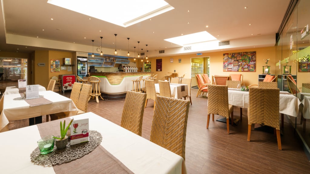 das Restaurant des Sportcenters Marco Polo mit beigen Flechtstühlen und weißen Tischtüchern mit hellbraunen Akzenten