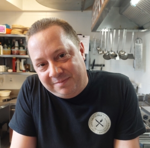 Restaurantleiter Paul Fritscher steht lächelnd in der Küche. Sein Kopf ist leicht geneigt. Auf seinem Shirt ist das Logo von "Paul Fritscher Gastronomie" zu sehen.